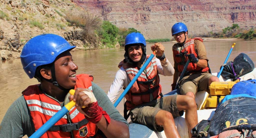 Three students wearing safety gear paddle a raft on calm water. 